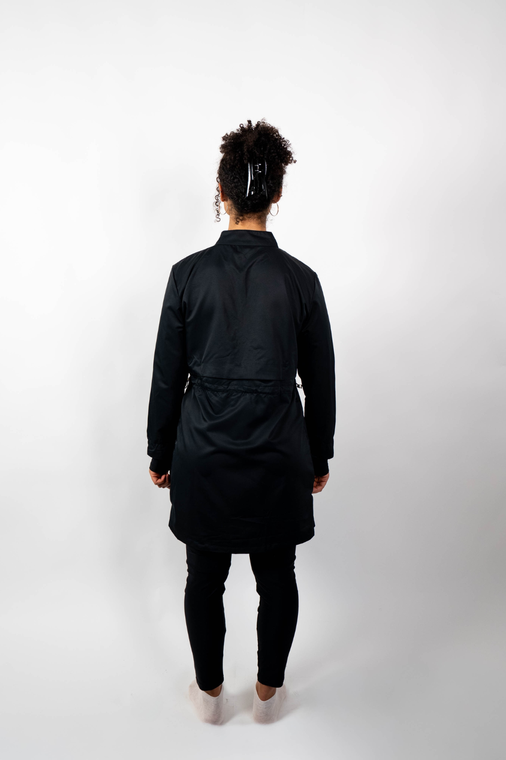 Rückenansicht einer Frau, die eine schwarze Sport-Tunika mit Stehkragen und einem geschichteten Look durch einen Taillenzug trägt.