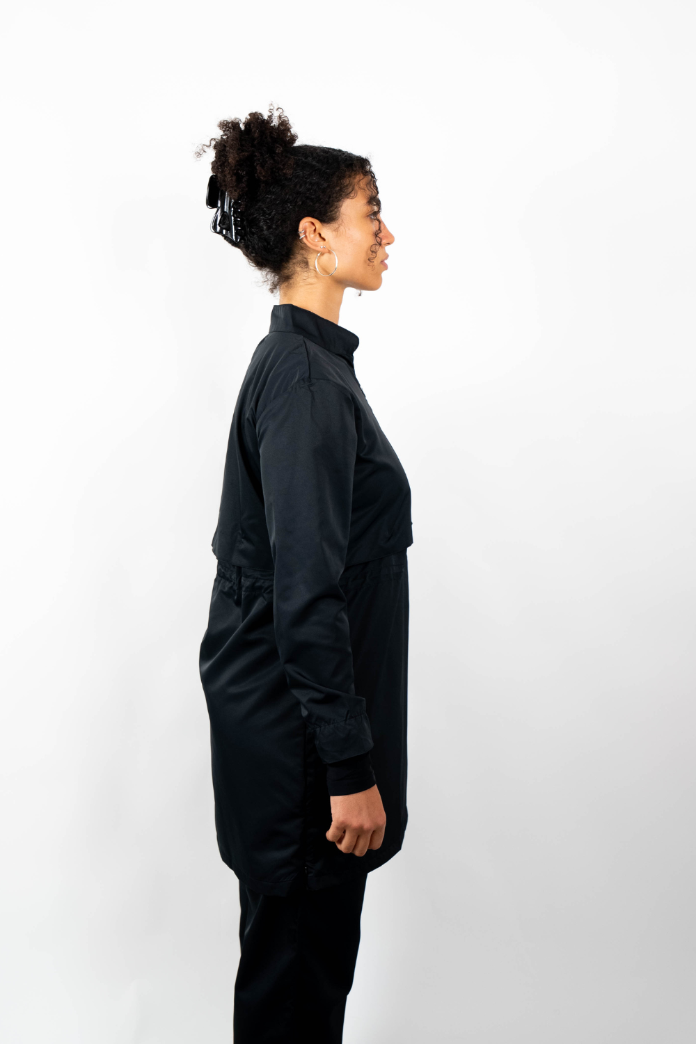 Seitenansicht einer Frau, die eine schwarze Sport-Tunika mit Stehkragen und einem geschichteten Look durch einen Taillenzug trägt.