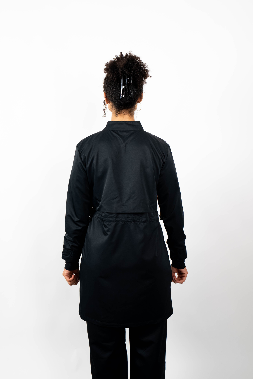 Rückenansicht einer Frau, die eine schwarze Sport-Tunika mit Stehkragen und einem geschichteten Look durch einen Taillenzug trägt.