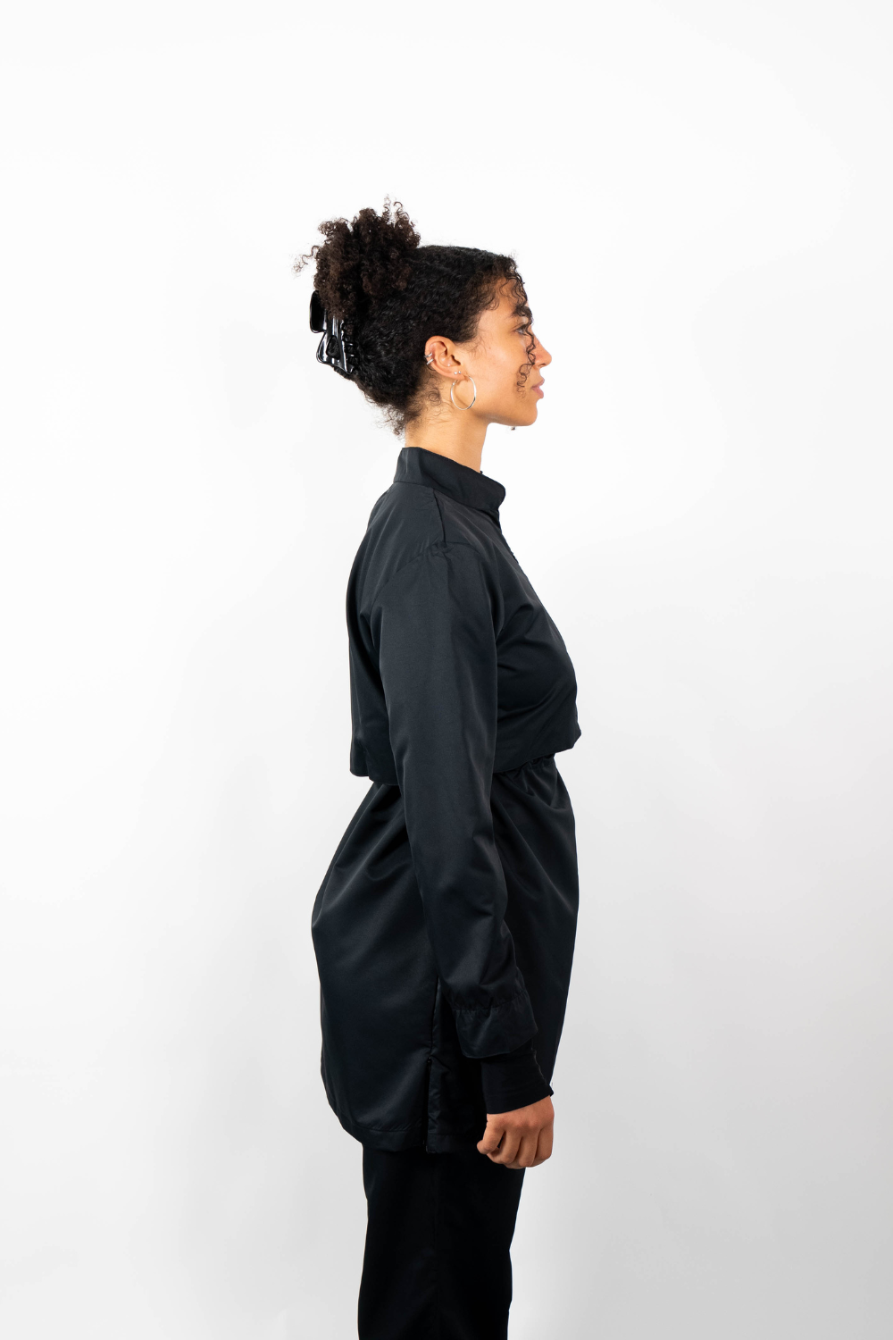Seitenansicht einer Frau, die eine schwarze Sport-Tunika mit Stehkragen und einem geschichteten Look durch einen Taillenzug trägt.