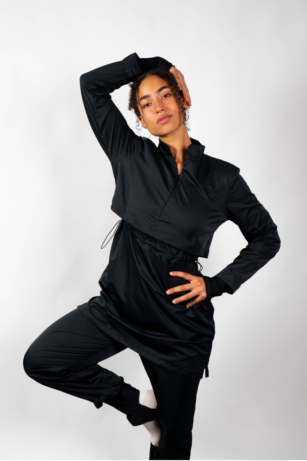 Ganzkörperaufnahme einer Frau in einem schwarzen Sport-Outfit (Tunika-Oberteil mit geschichtetem Look und Sporthose) in einer Balance-Pose.