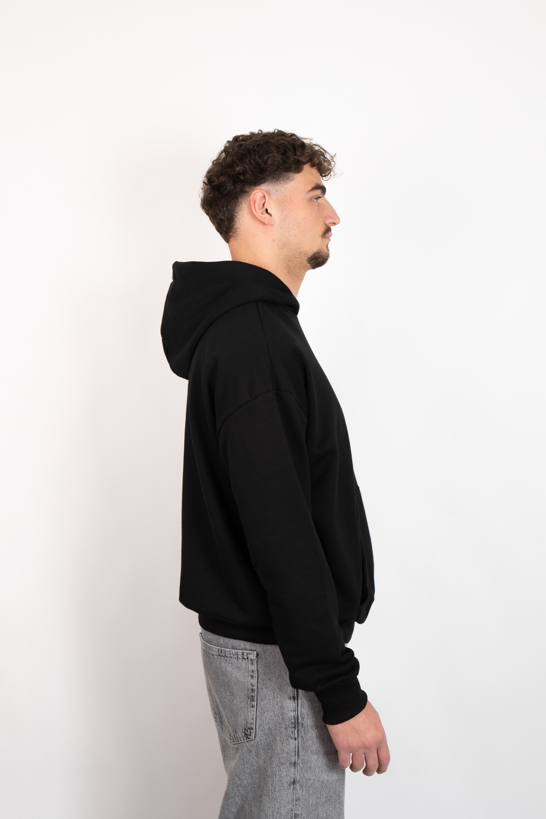 Seitenansicht eines Mannes, der einen schwarzen Oversize Hoodie mit Kapuze trägt, kombiniert mit grauen Jeans.