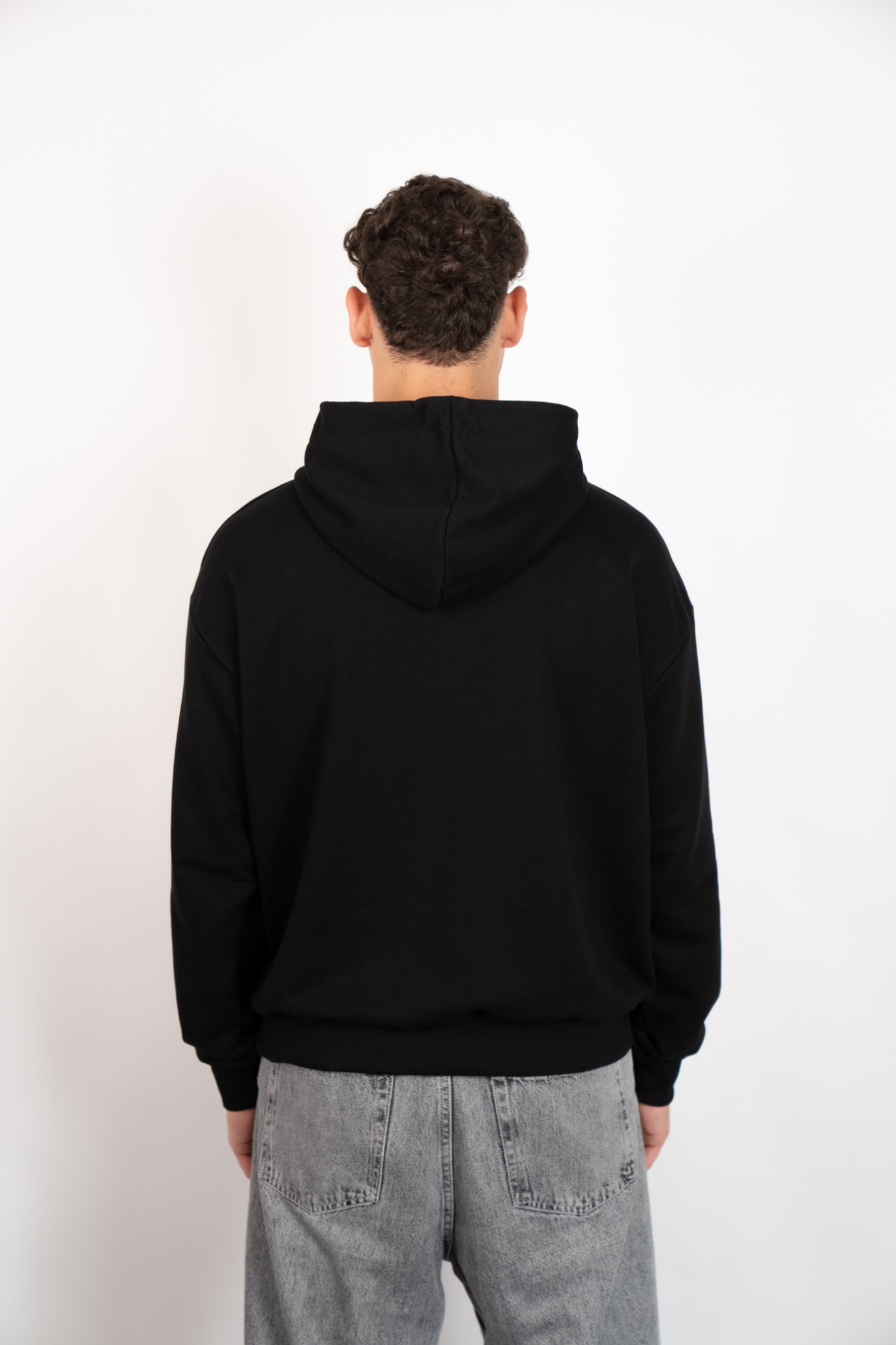 Rückenansicht eines Mannes, der einen schwarzen Oversize Hoodie mit Kapuze trägt, kombiniert mit grauen Jeans.