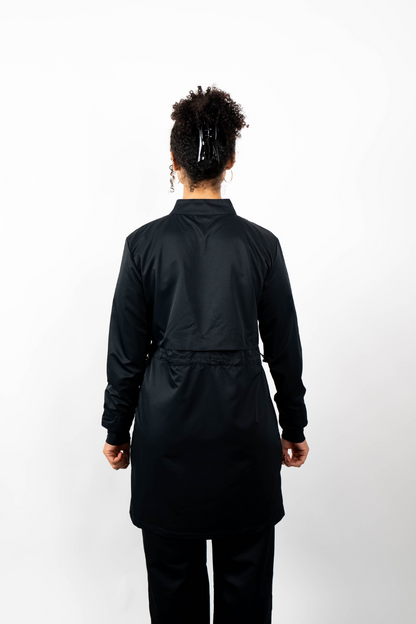 Rückenansicht einer Frau, die eine schwarze Sport-Tunika mit Stehkragen und einem geschichteten Look durch einen Taillenzug trägt.