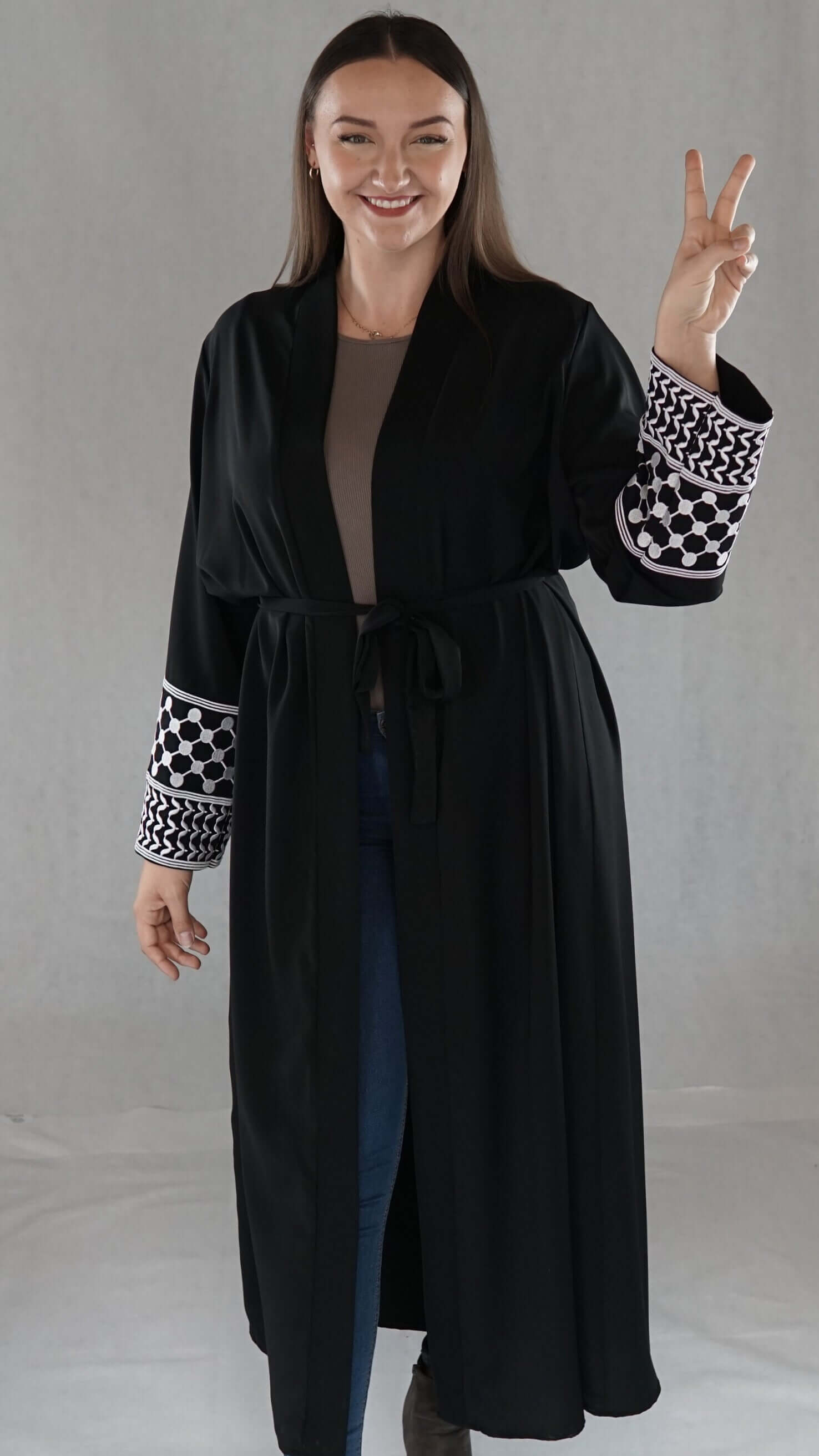 Schwarze Abaya mit palästinensischem Design, elegant und traditionell, präsentiert von Model mit friedlichem Zeichen.