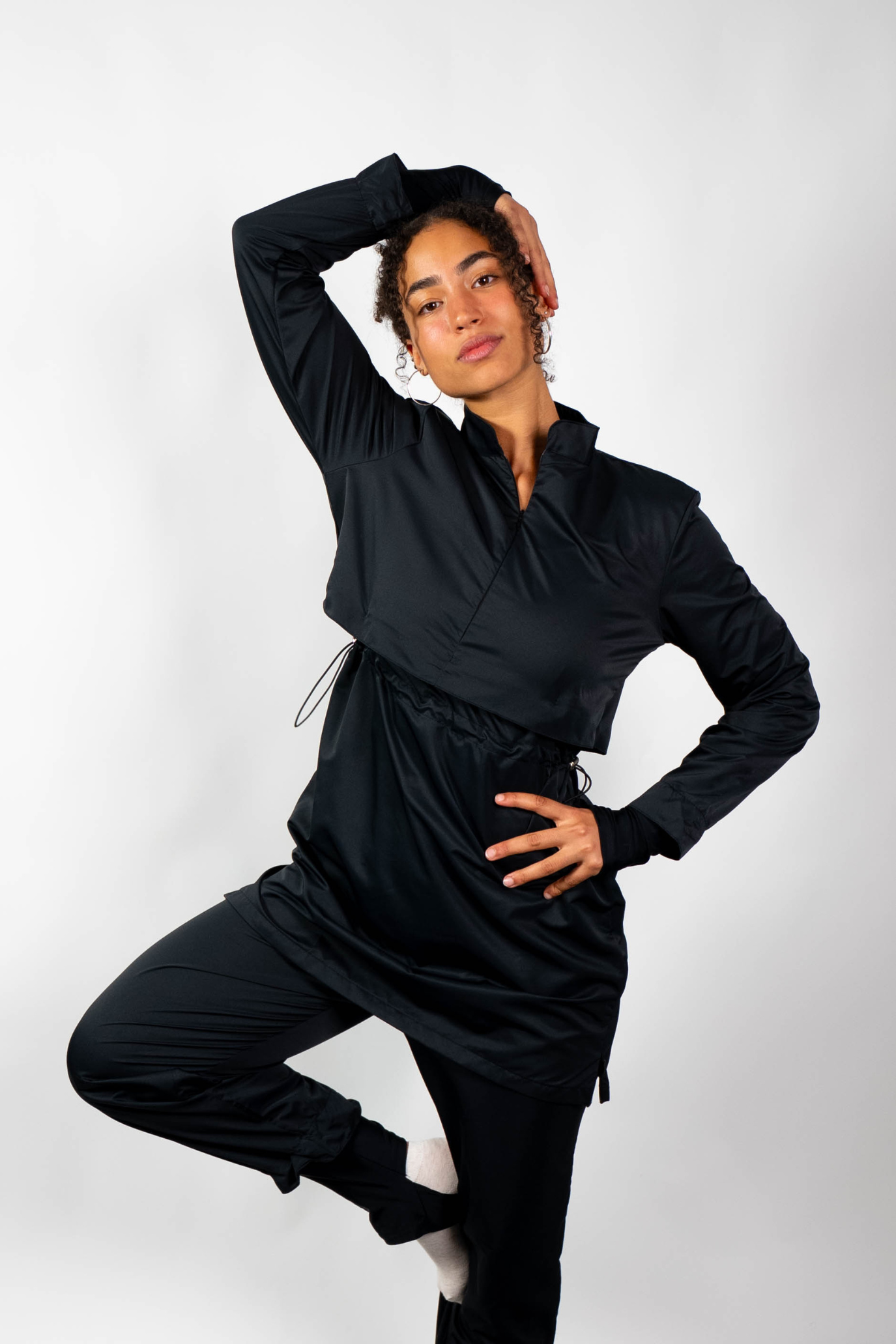 Ganzkörperaufnahme einer Frau in einem schwarzen, geschichteten Bade-Outfit (lange Tunika und Sporthose) in einer dynamischen Pose.