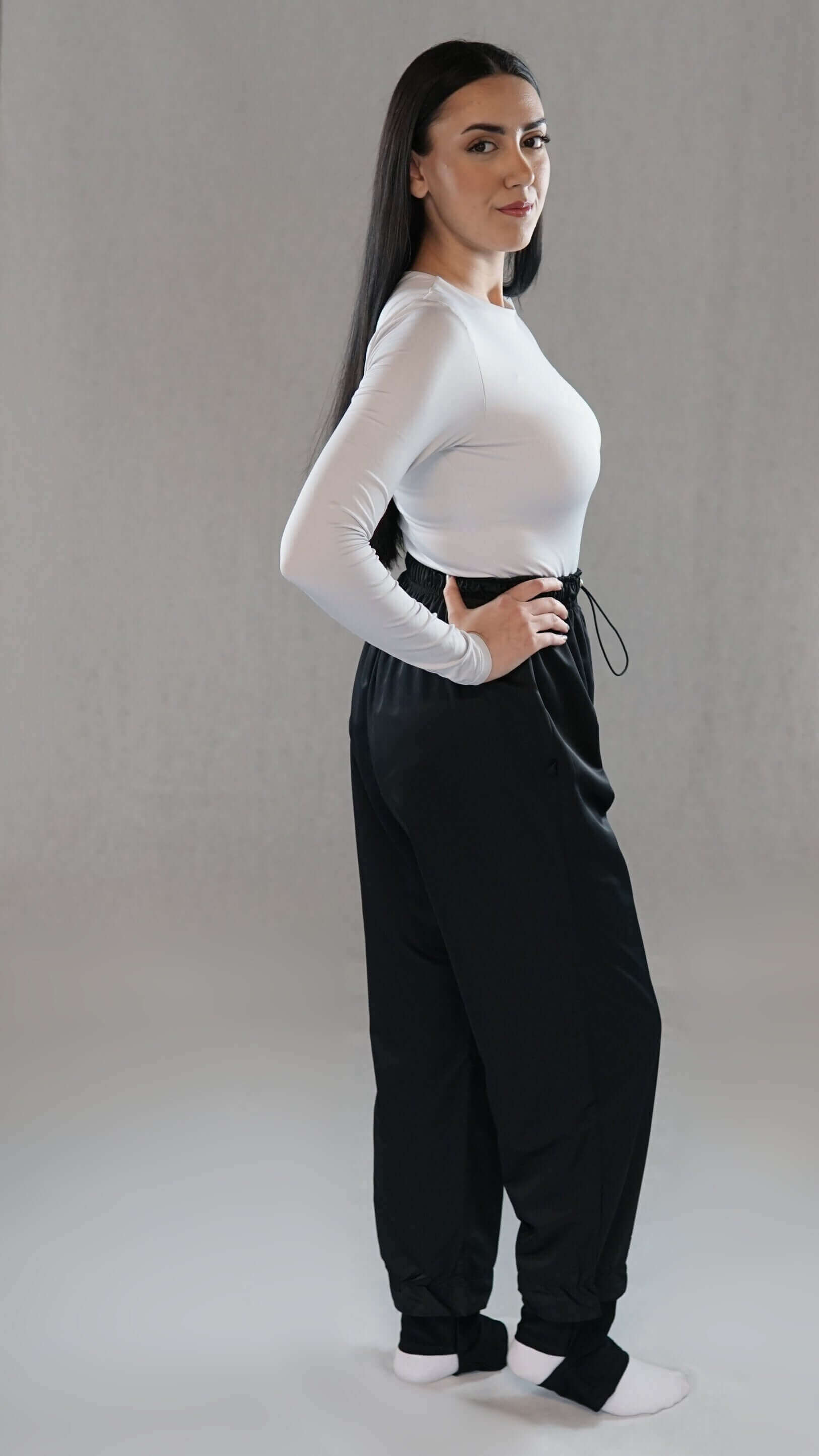 Model wearing a long-sleeve white top and black sport pants, posing confidently in a minimalist studio.