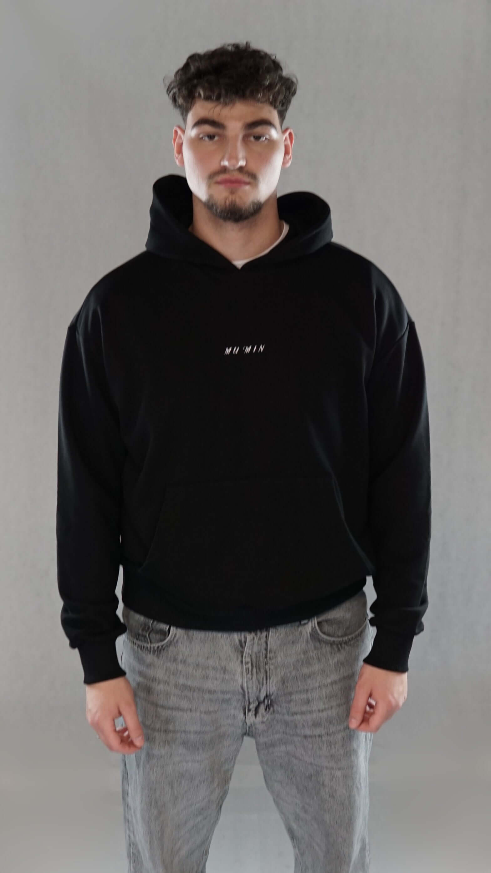 Man wearing black oversize hoodie with minimalist "Mu’min" embroidery on chest, symbolizing spirituality and inner faith.