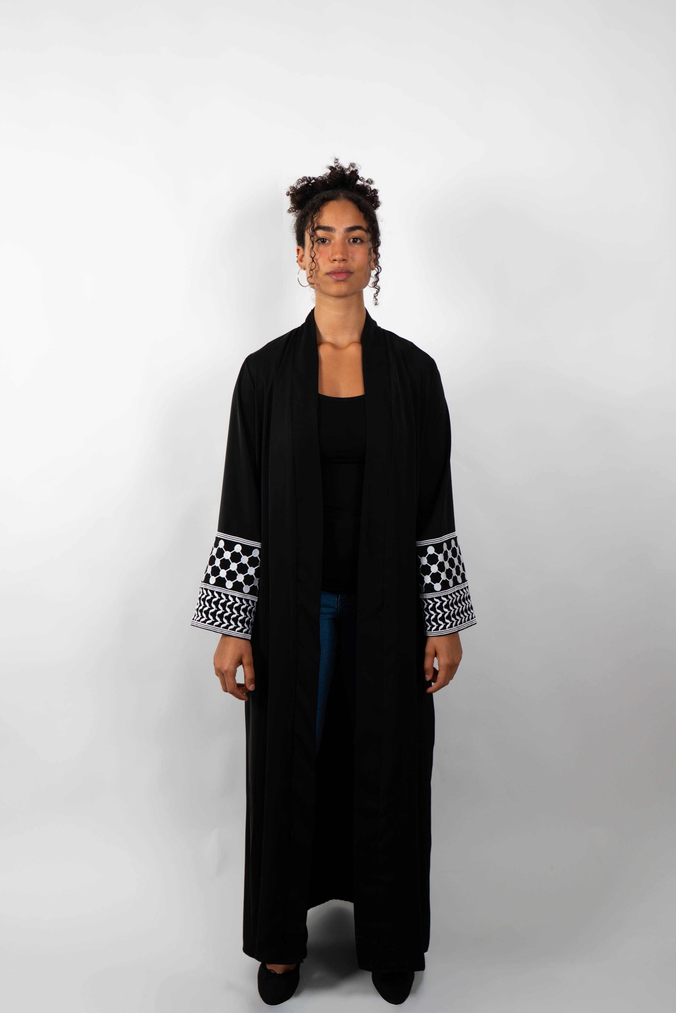 Frontalansicht einer Frau, die eine lange, schwarze Abaya trägt. Die Abaya hat weite Ärmel, deren Enden mit einem schwarz-weißen Muster verziert sind.