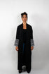 Frontalansicht einer Frau, die eine lange, schwarze Abaya trägt. Die Abaya hat weite Ärmel, deren Enden mit einem schwarz-weißen Muster verziert sind.