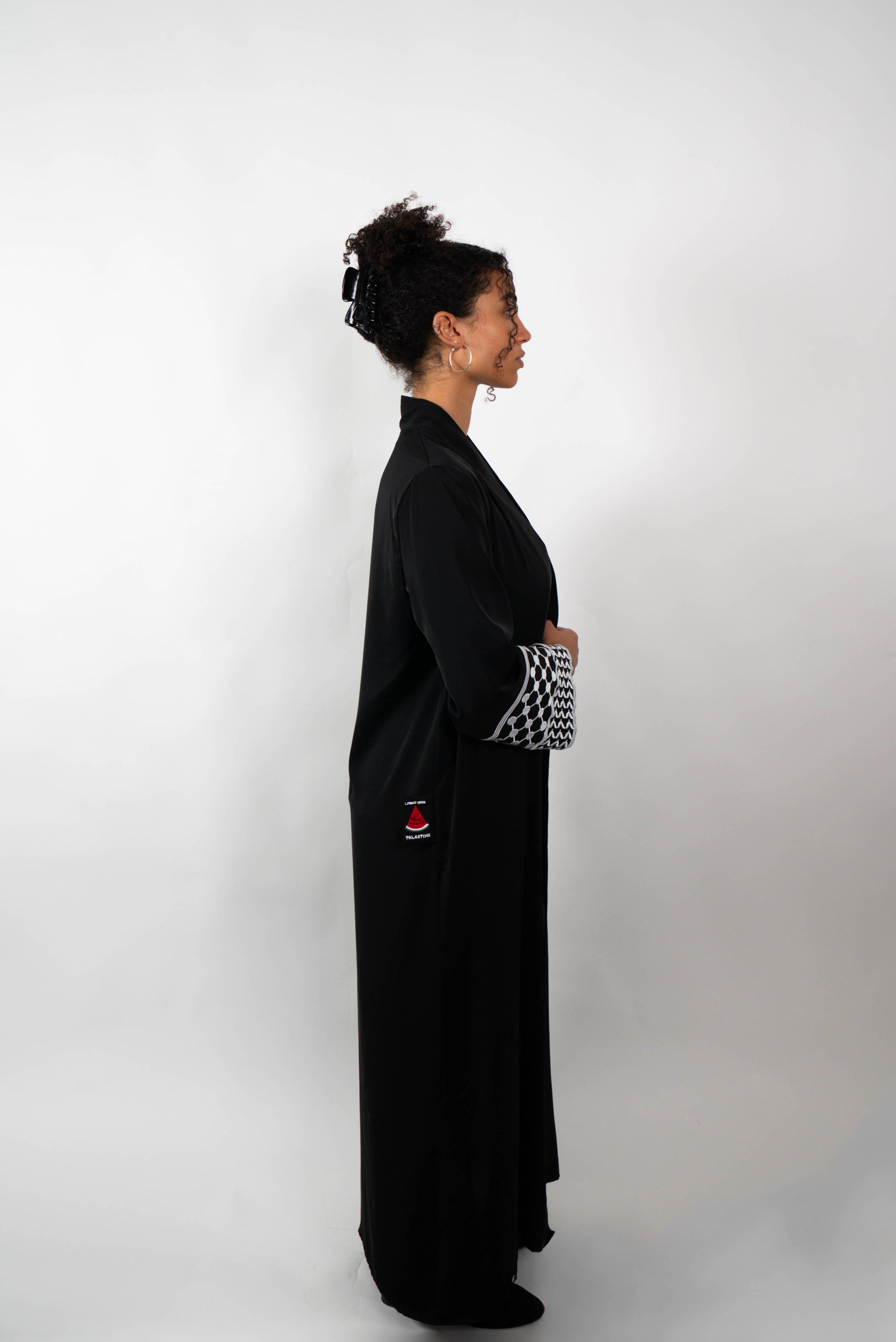Seitenansicht einer Frau, die eine lange, schwarze Abaya trägt. Die Abaya hat weite Ärmel, deren Enden mit einem schwarz-weißen Muster verziert sind.