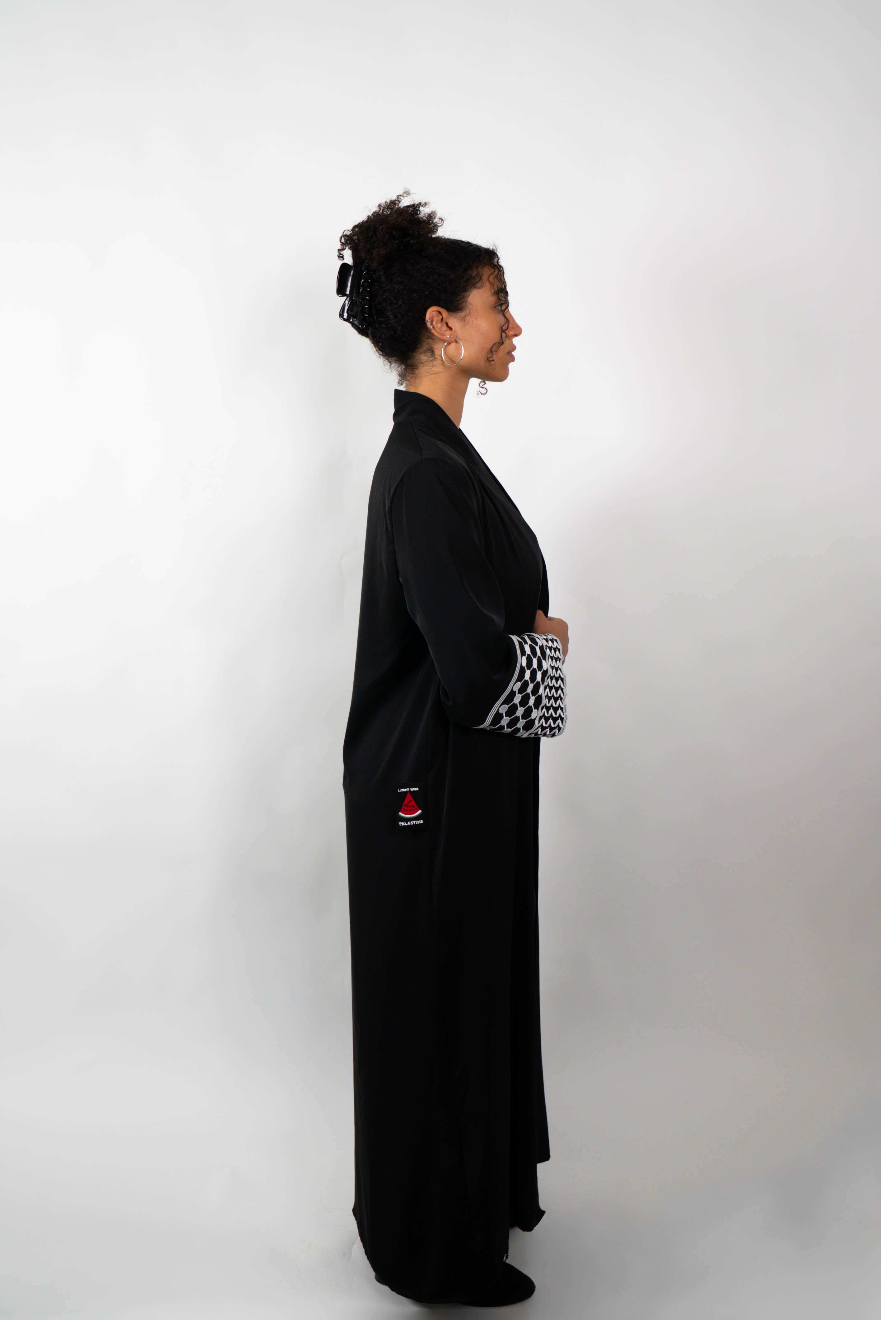 Seitenansicht einer Frau, die eine lange, schwarze Abaya trägt. Die Abaya hat weite Ärmel, deren Enden mit einem schwarz-weißen Muster verziert sind.