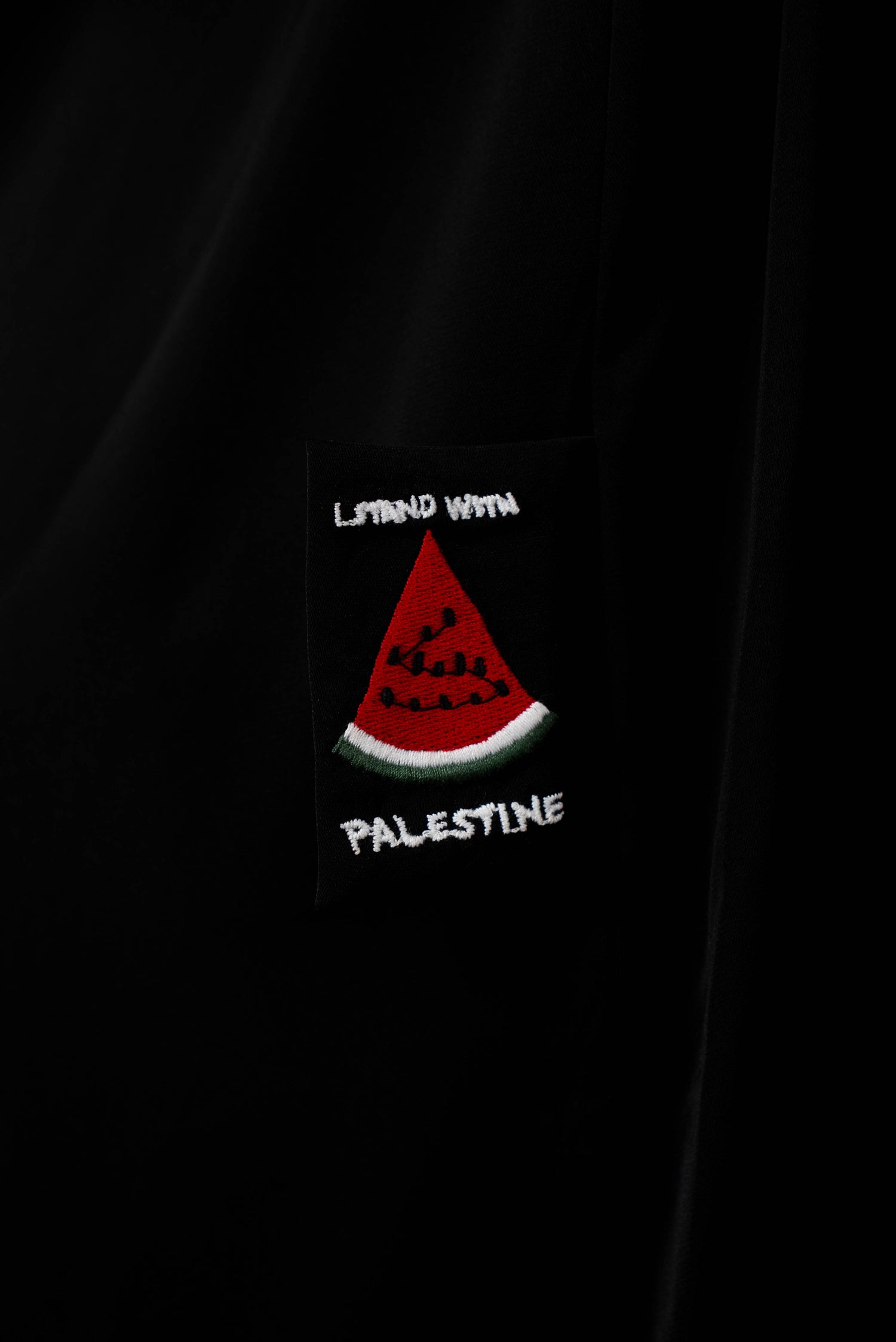 Nahaufnahme auf das Detail der schwarzen Abaya, die einen gestickten, schwarzen Patch mit der Aufschrift &quot;STAND WITH PALESTINE&quot; und einer roten Wassermelone zeigt.