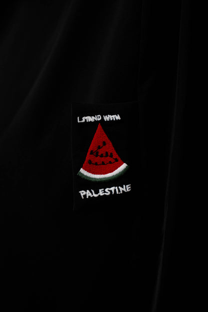 Nahaufnahme auf das Detail der schwarzen Abaya, die einen gestickten, schwarzen Patch mit der Aufschrift &quot;STAND WITH PALESTINE&quot; und einer roten Wassermelone zeigt.
