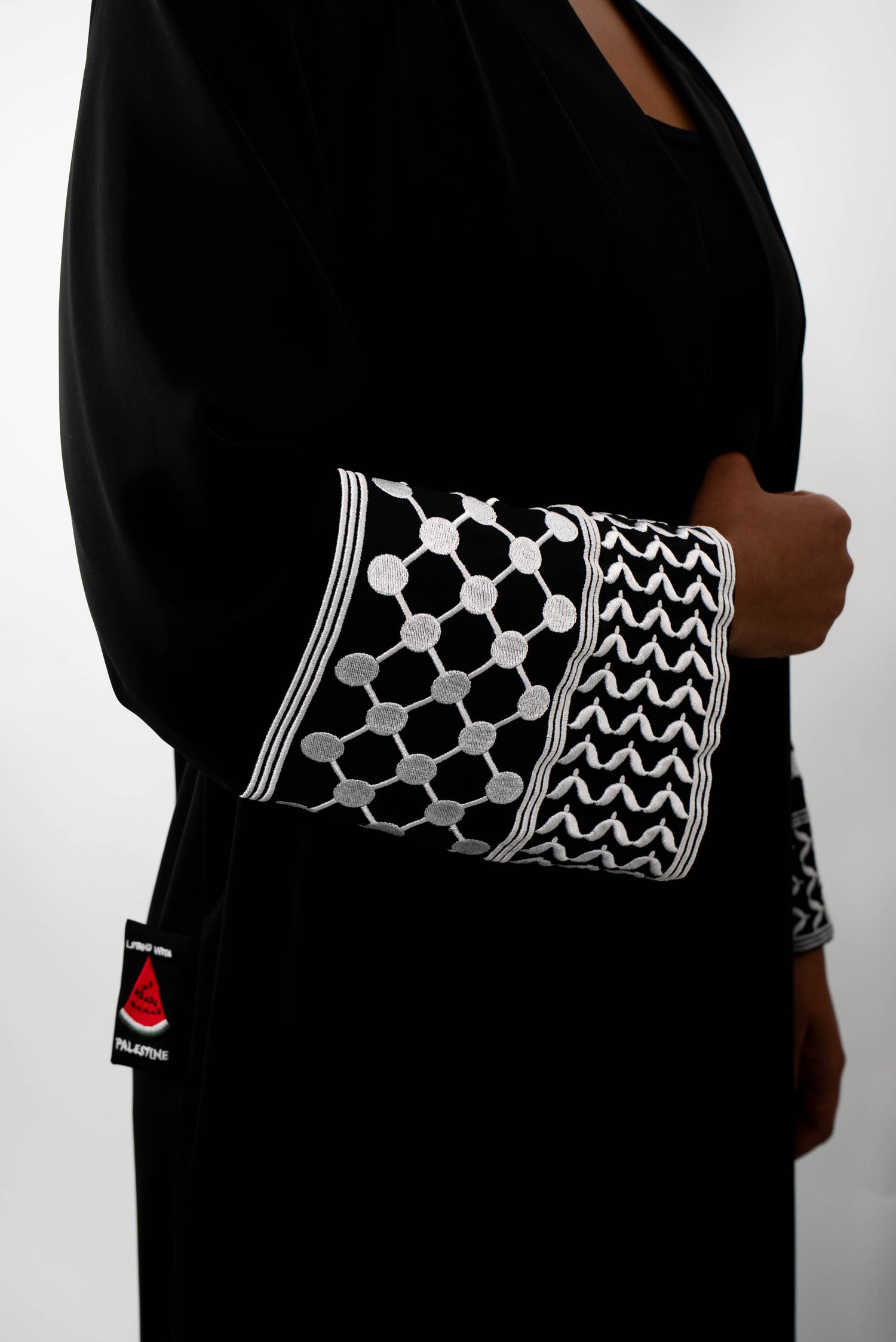 Nahaufnahme des Ärmelabschlusses einer schwarzen Abaya. Der Ärmel ist mit detailreicher, weißer Stickerei in einem geometrischen, netzartigen Muster und einem Zickzack-Muster verziert. Auch der schwarze &quot;STAND WITH PALESTINE&quot;-Patch ist sichtbar.