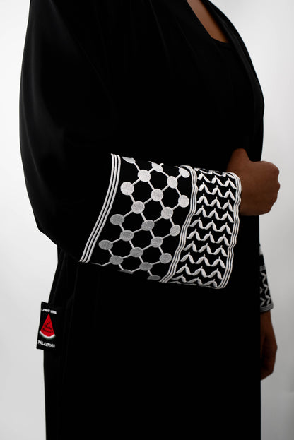 Nahaufnahme des Ärmelabschlusses einer schwarzen Abaya. Der Ärmel ist mit detailreicher, weißer Stickerei in einem geometrischen, netzartigen Muster und einem Zickzack-Muster verziert. Auch der schwarze &quot;STAND WITH PALESTINE&quot;-Patch ist sichtbar.