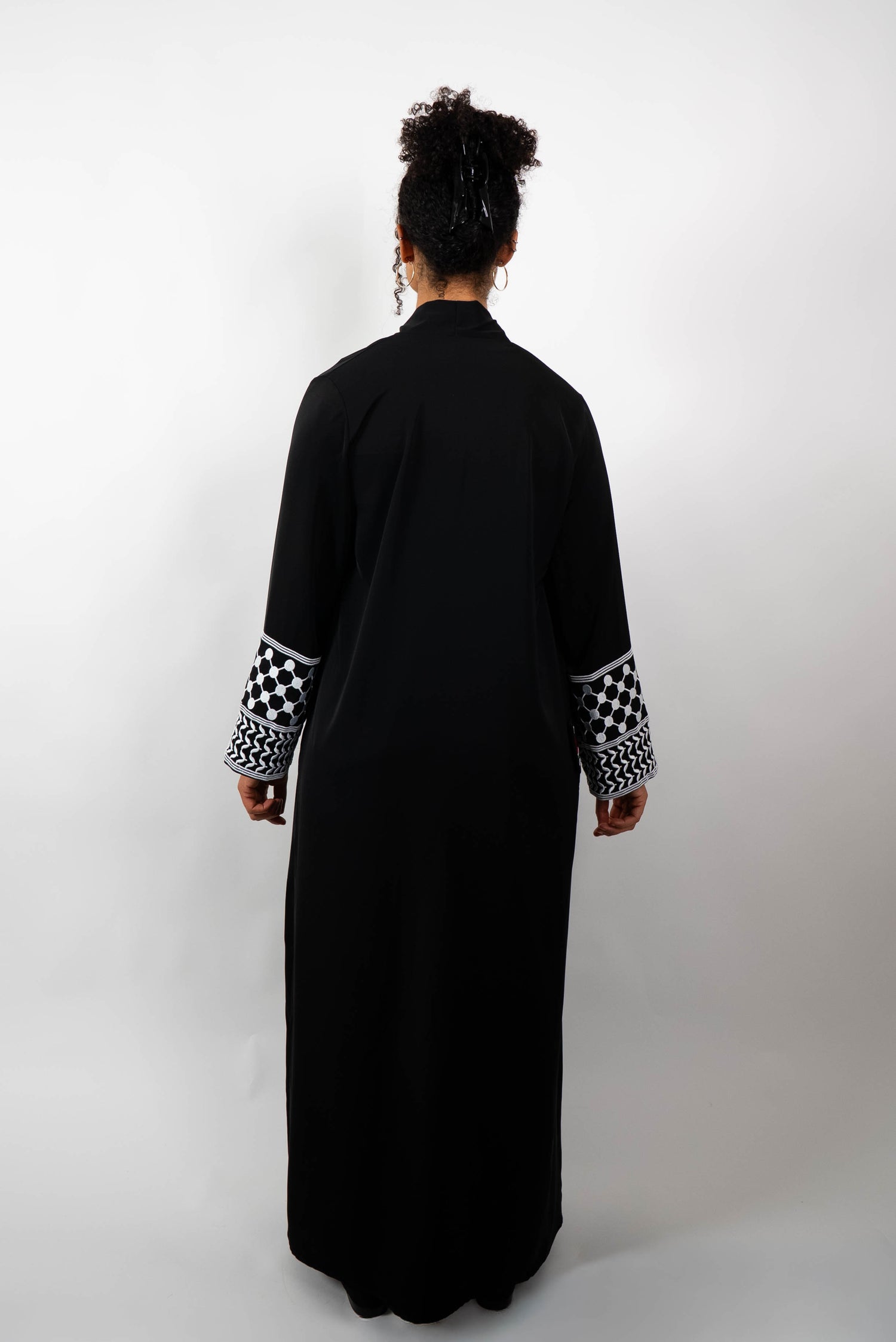 Rückenansicht einer Frau in einer langen, schwarzen Abaya. Die Ärmelabschlüsse sind mit schwarz-weißen Kufiya-ähnlichen Mustern verziert.