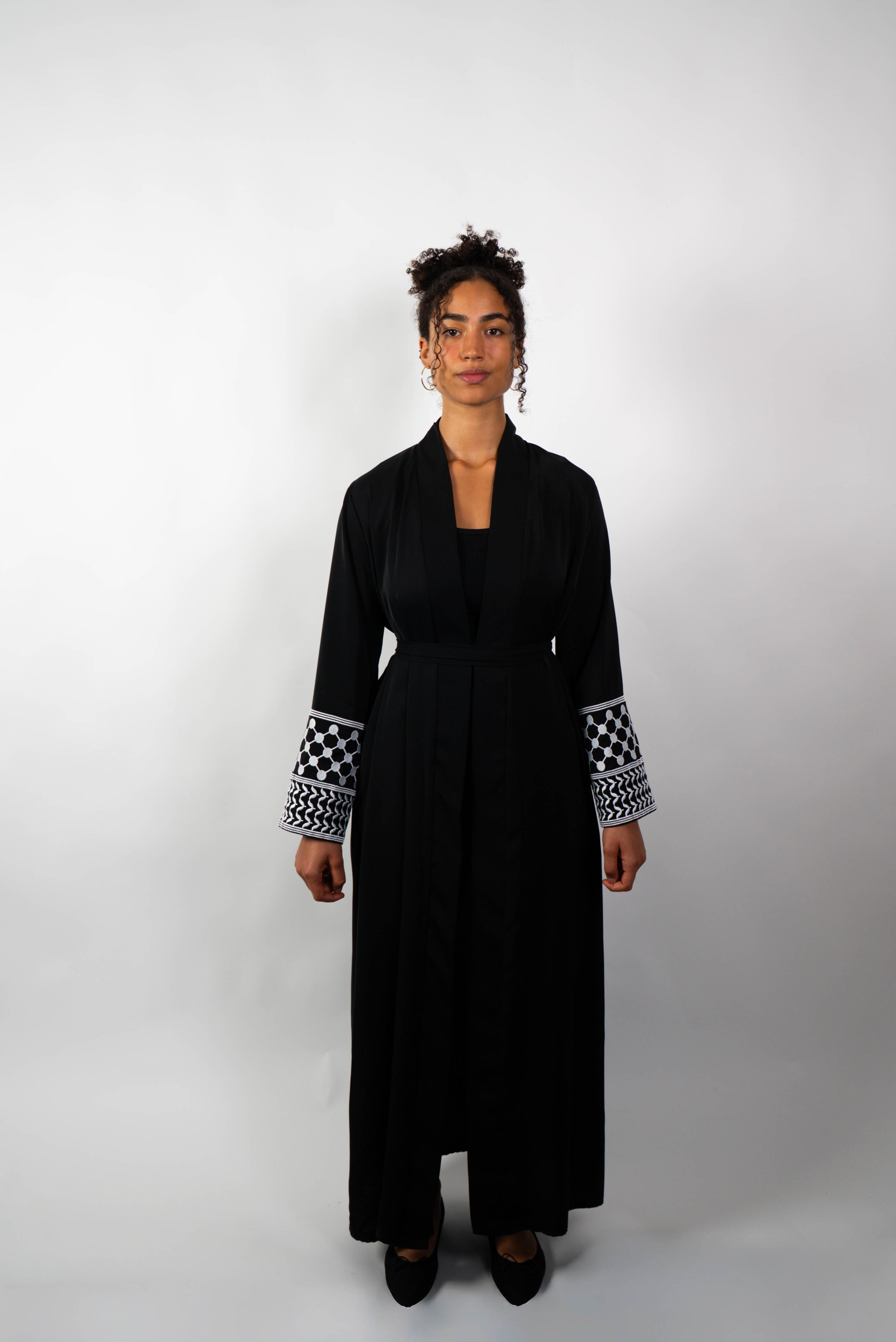 Frontalansicht einer Frau in einer langen, schwarzen Abaya, die locker gebunden ist. Die Ärmelabschlüsse sind mit schwarz-weißen Kufiya-ähnlichen Mustern verziert.