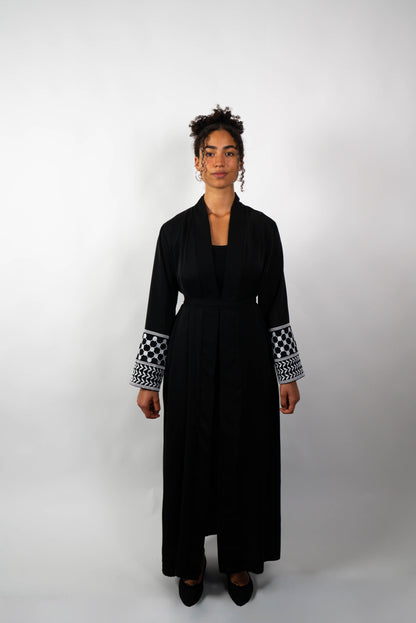Frontalansicht einer Frau in einer langen, schwarzen Abaya, die locker gebunden ist. Die Ärmelabschlüsse sind mit schwarz-weißen Kufiya-ähnlichen Mustern verziert.