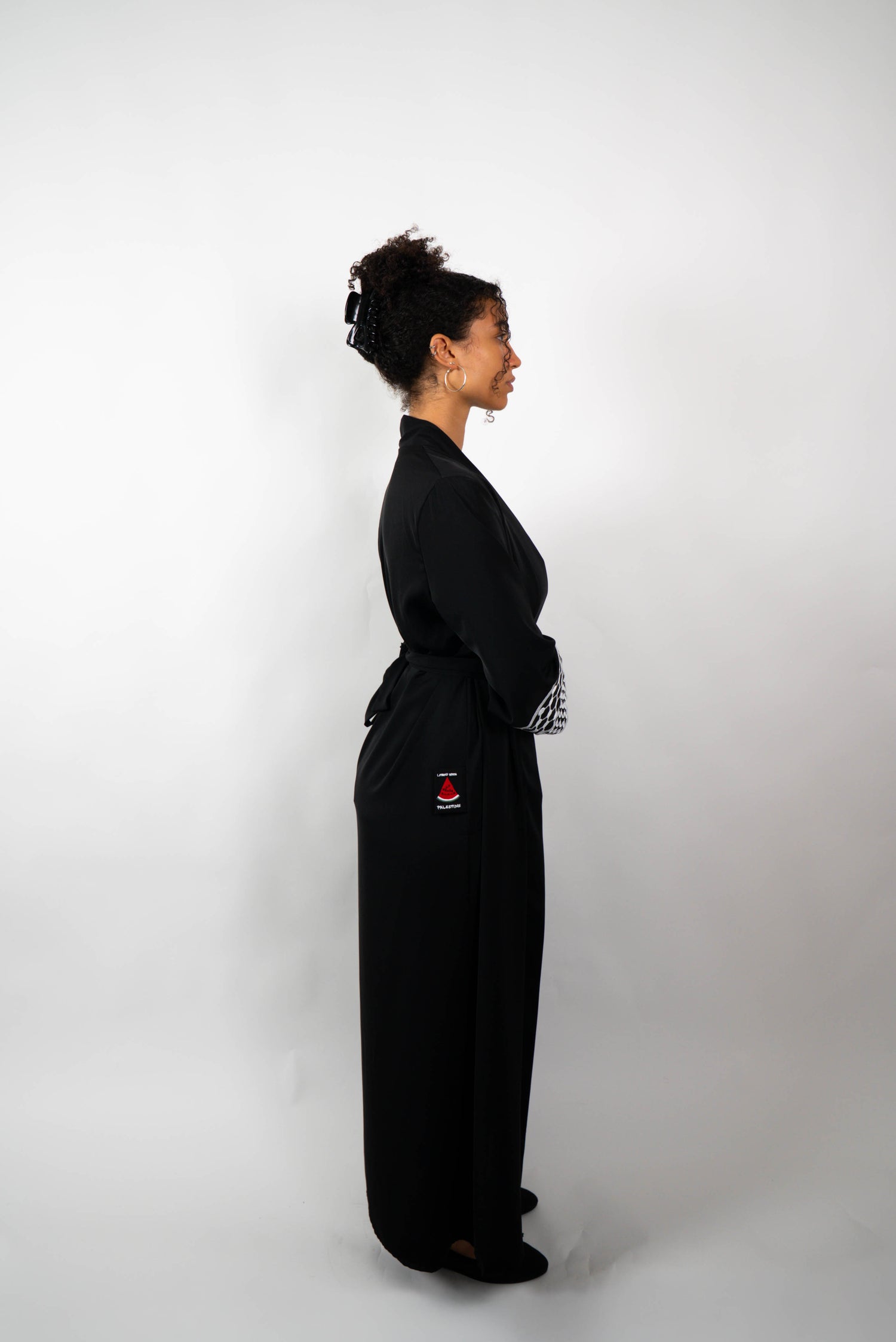 Seitenansicht einer Frau in einer langen, schwarzen Abaya, die locker gebunden ist. Die Ärmelabschlüsse sind mit schwarz-weißen Kufiya-ähnlichen Mustern verziert.