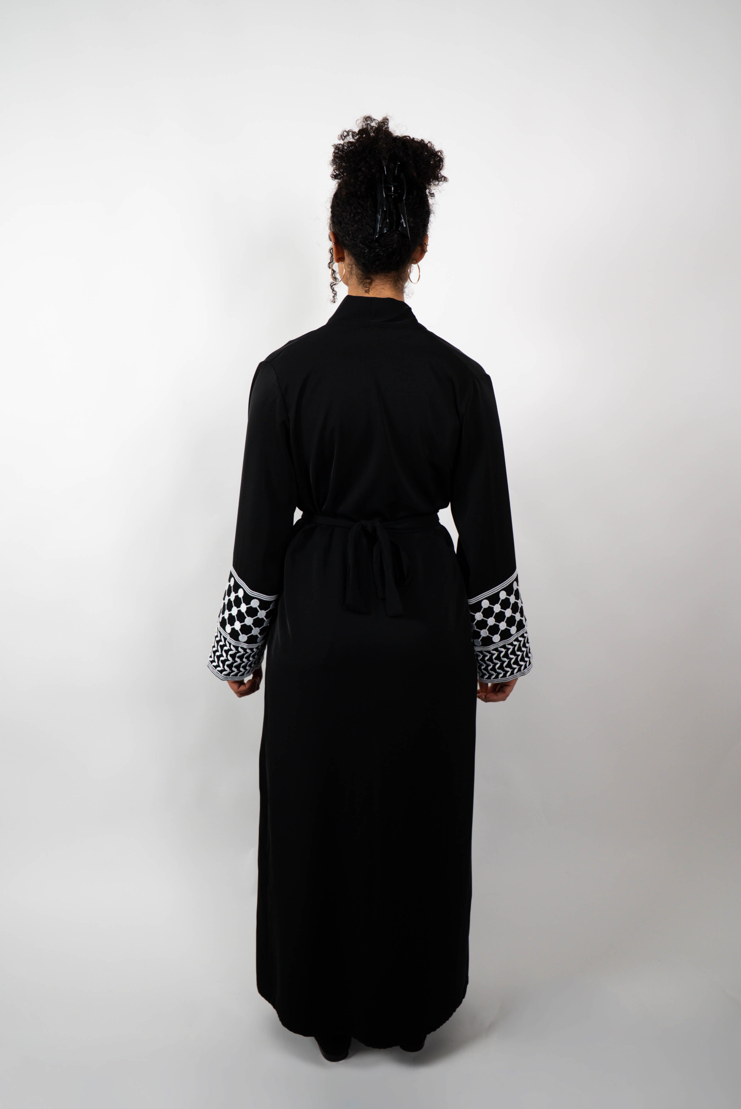 Rückenansicht einer Frau in einer langen, schwarzen Abaya, die mit einem Gürtel im Rücken gebunden ist. Die Ärmelabschlüsse sind mit schwarz-weißen Kufiya-ähnlichen Mustern verziert.
