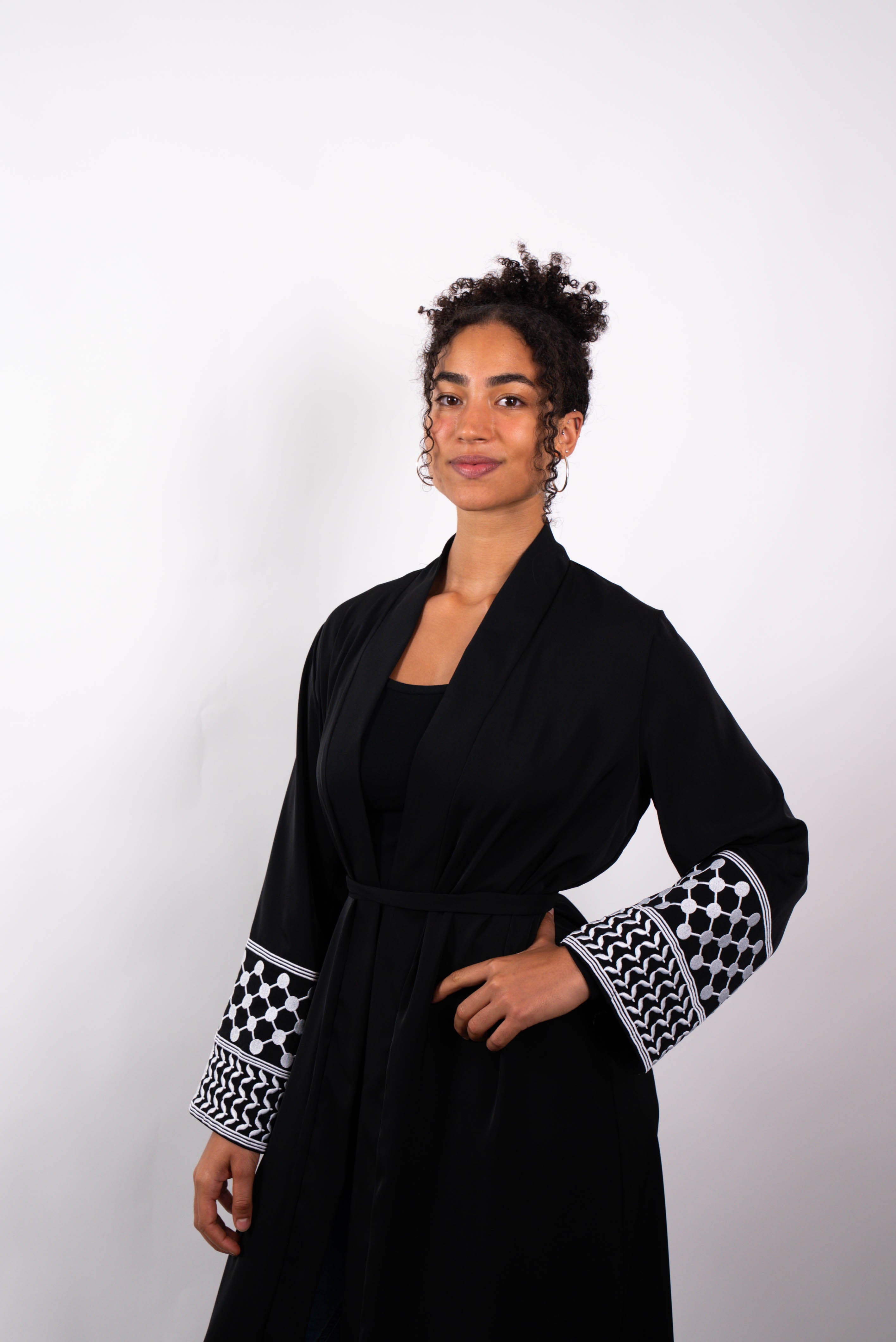 Halbkörper-Aufnahme einer lächelnden Frau in einer langen, schwarzen Abaya mit Gürtel. Die weiten Ärmel sind mit schwarz-weißen Kufiya-ähnlichen Mustern verziert.