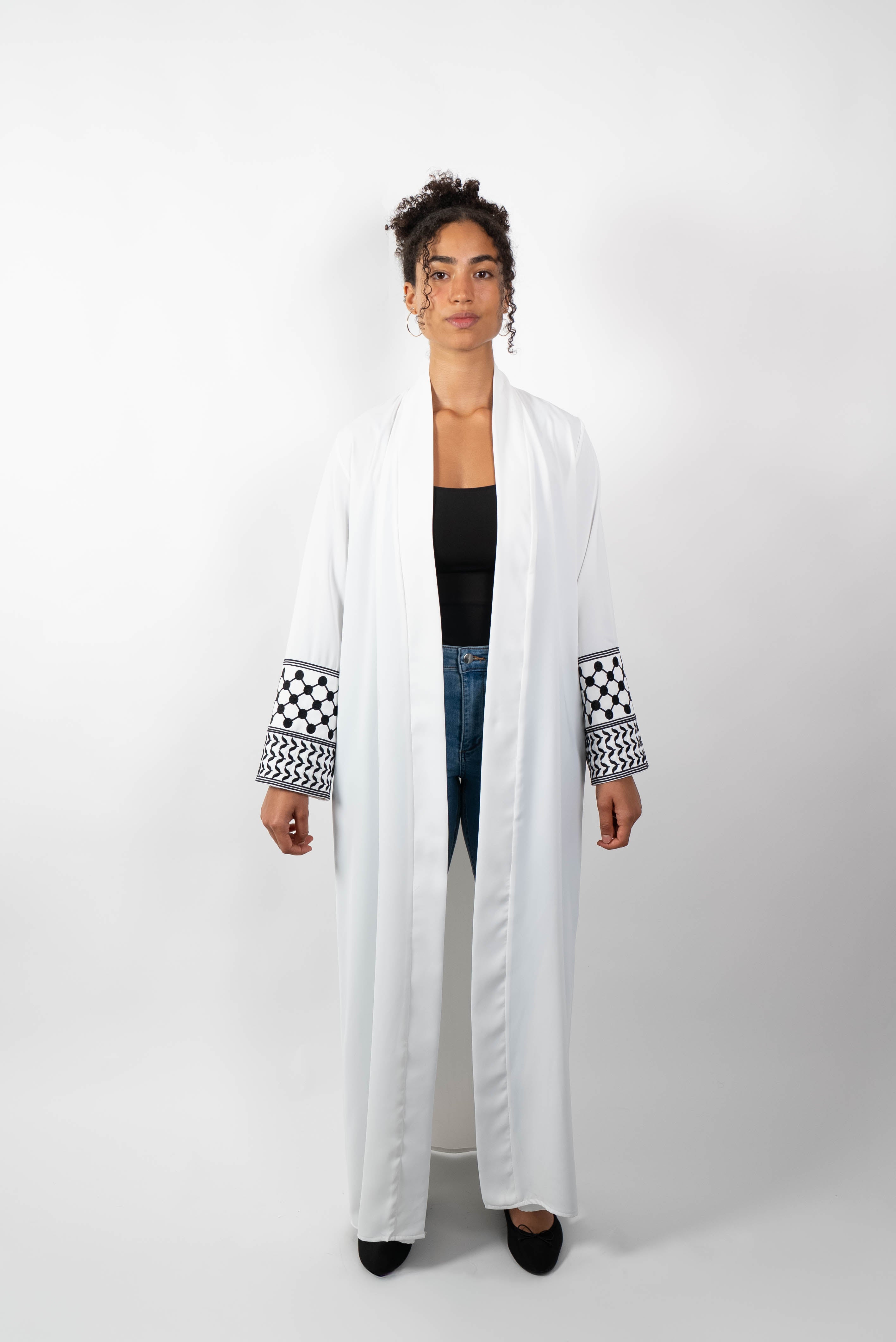 Frontalansicht einer Frau, die eine lange, offene, weiße Abaya trägt. Die weiten Ärmel sind mit einem auffälligen schwarz-weißen Muster, das Kufiya-ähnlichen Mustern nachempfunden ist, verziert.
