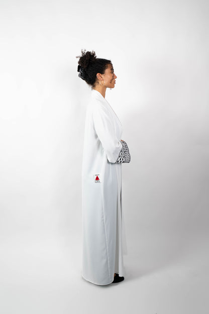 Seitenansicht einer Frau, die eine lange, offene, weiße Abaya trägt. Die weiten Ärmel sind mit einem auffälligen schwarz-weißen Muster, das Kufiya-ähnlichen Mustern nachempfunden ist, verziert.