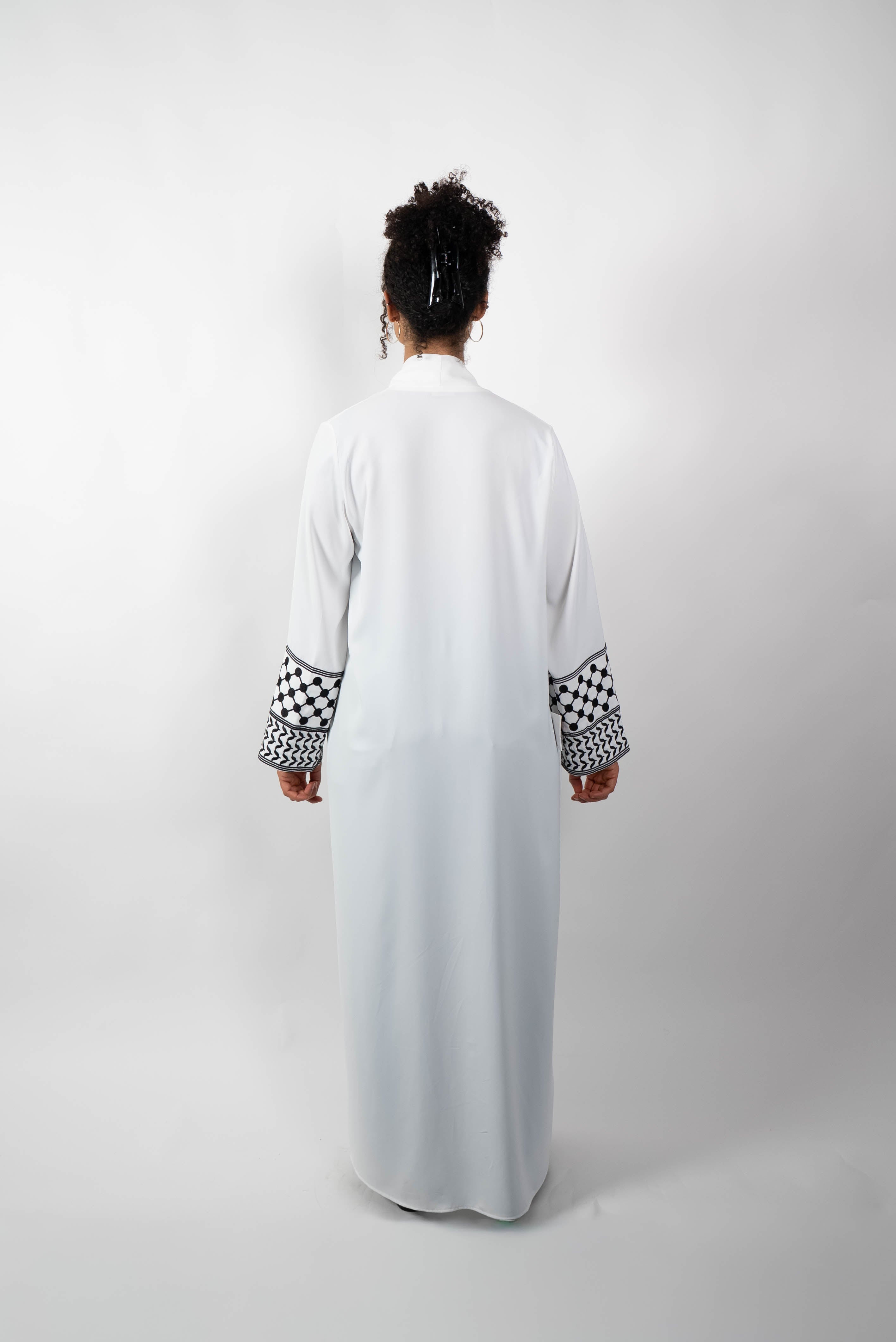 Rückenansicht einer Frau, die eine lange, offene, weiße Abaya trägt. Die weiten Ärmel sind mit einem auffälligen schwarz-weißen Muster, das Kufiya-ähnlichen Mustern nachempfunden ist, verziert.