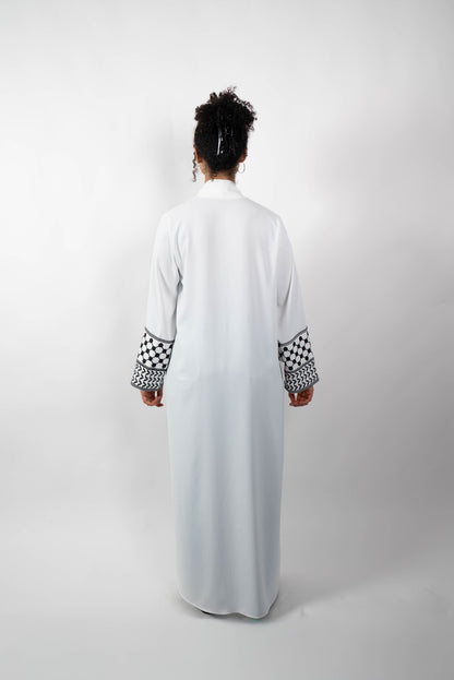 Rückenansicht einer Frau, die eine lange, offene, weiße Abaya trägt. Die weiten Ärmel sind mit einem auffälligen schwarz-weißen Muster, das Kufiya-ähnlichen Mustern nachempfunden ist, verziert.