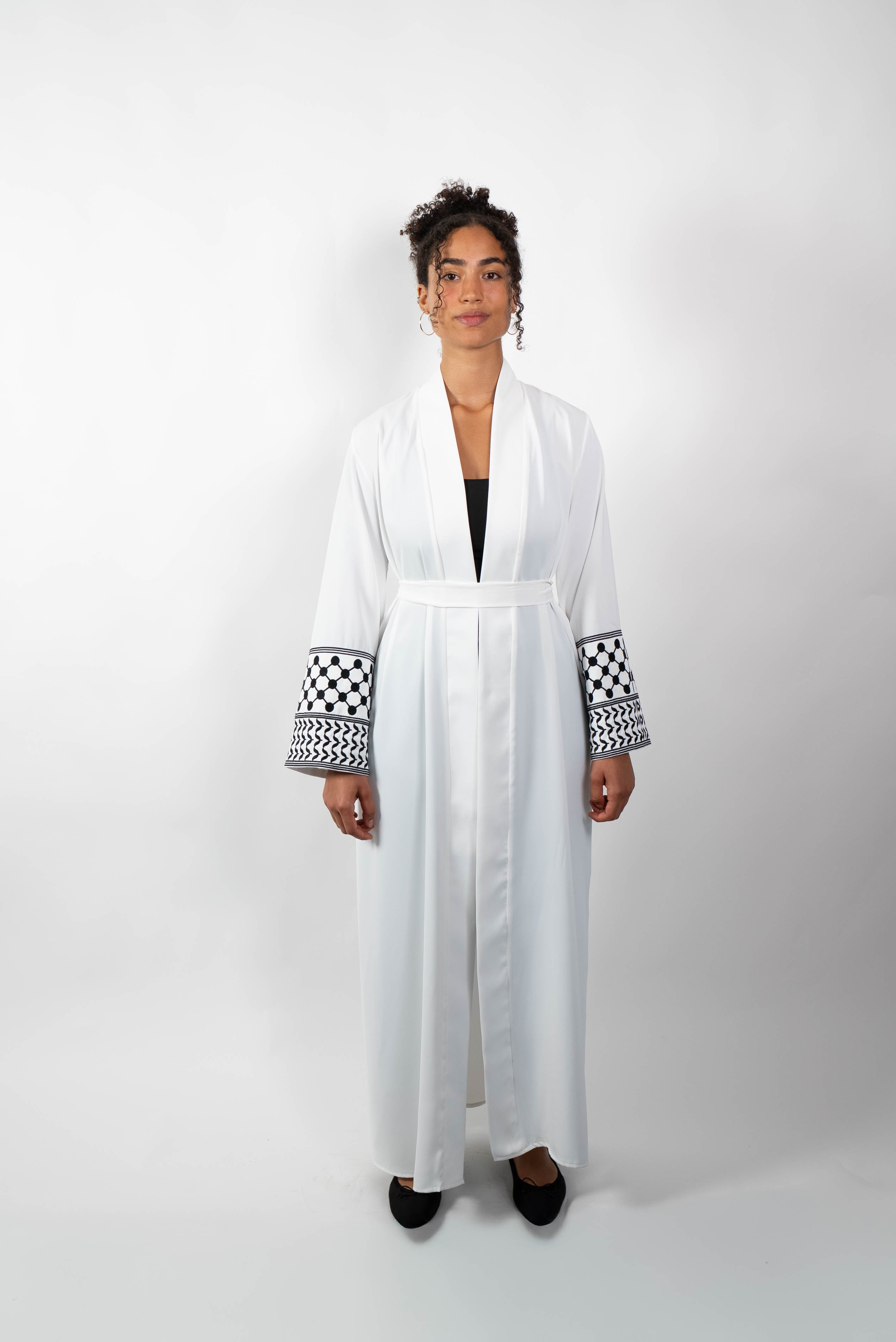 Frontalansicht einer Frau in einer langen, weißen Abaya, die mit einem Stoffgürtel geschlossen ist. Die weiten Ärmel sind mit einem auffälligen schwarz-weißen Muster, das Kufiya-ähnlichen Mustern nachempfunden ist, verziert.