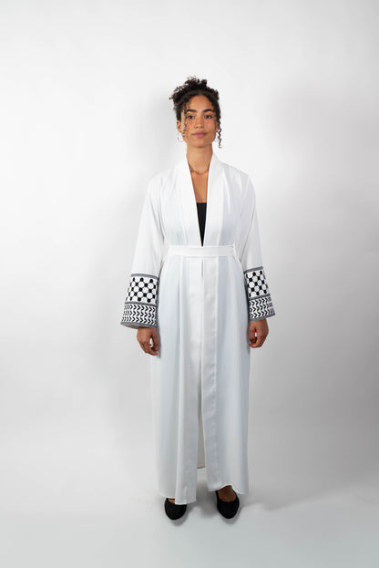 Frontalansicht einer Frau in einer langen, weißen Abaya, die mit einem Stoffgürtel geschlossen ist. Die weiten Ärmel sind mit einem auffälligen schwarz-weißen Muster, das Kufiya-ähnlichen Mustern nachempfunden ist, verziert.