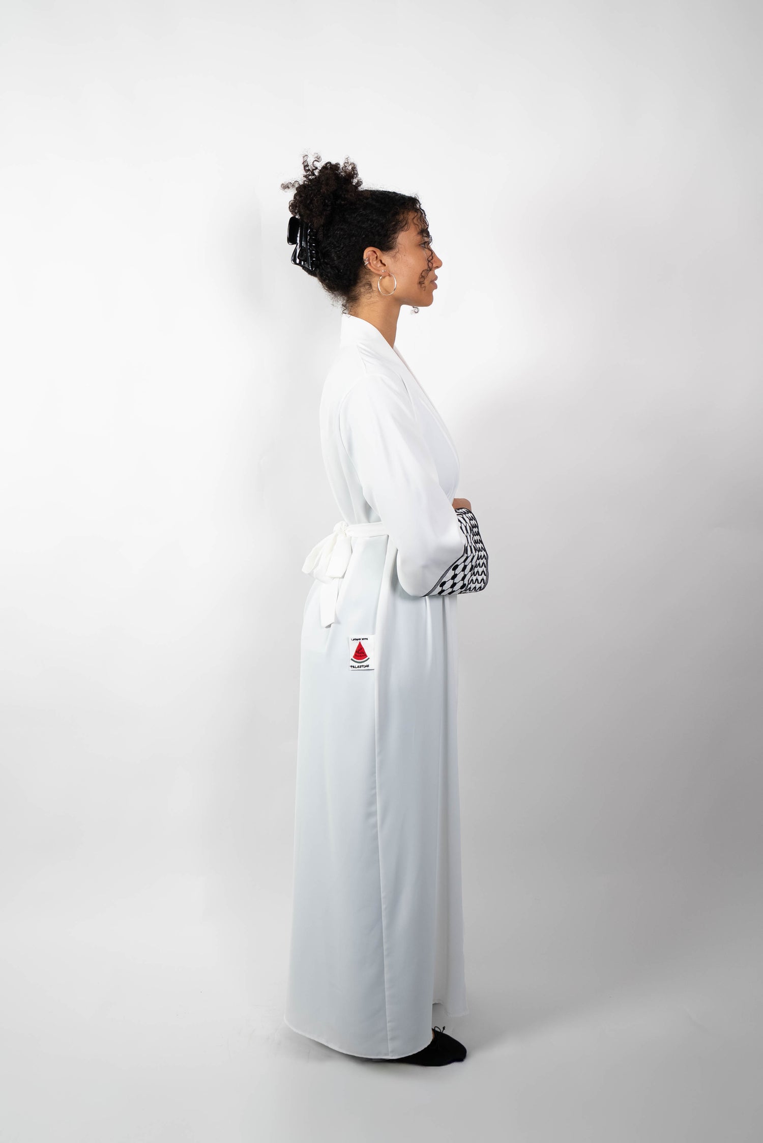 Seitenansicht einer Frau in einer langen, weißen Abaya, die mit einem Stoffgürtel geschlossen ist. Die weiten Ärmel sind mit einem auffälligen schwarz-weißen Muster, das Kufiya-ähnlichen Mustern nachempfunden ist, verziert.