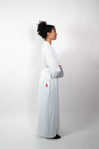 Seitenansicht einer Frau in einer langen, weißen Abaya, die mit einem Stoffgürtel geschlossen ist. Die weiten Ärmel sind mit einem auffälligen schwarz-weißen Muster, das Kufiya-ähnlichen Mustern nachempfunden ist, verziert.