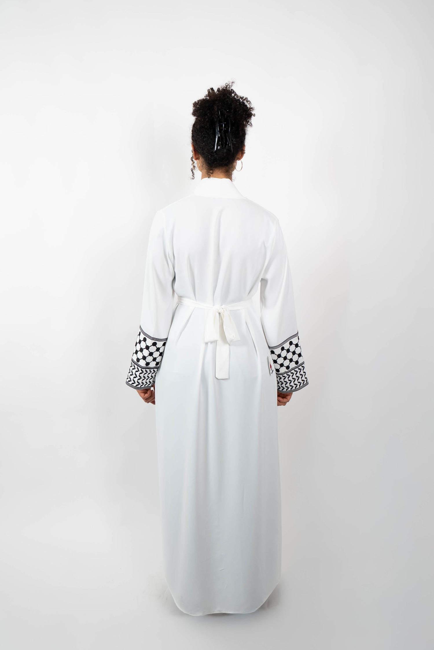 Rückenansicht einer Frau in einer langen, weißen Abaya, die mit einem Gürtel im Rücken geschlossen ist. Die Ärmelabschlüsse sind mit einem auffälligen schwarz-weißen Muster, das Kufiya-ähnlichen Mustern nachempfunden ist, verziert.