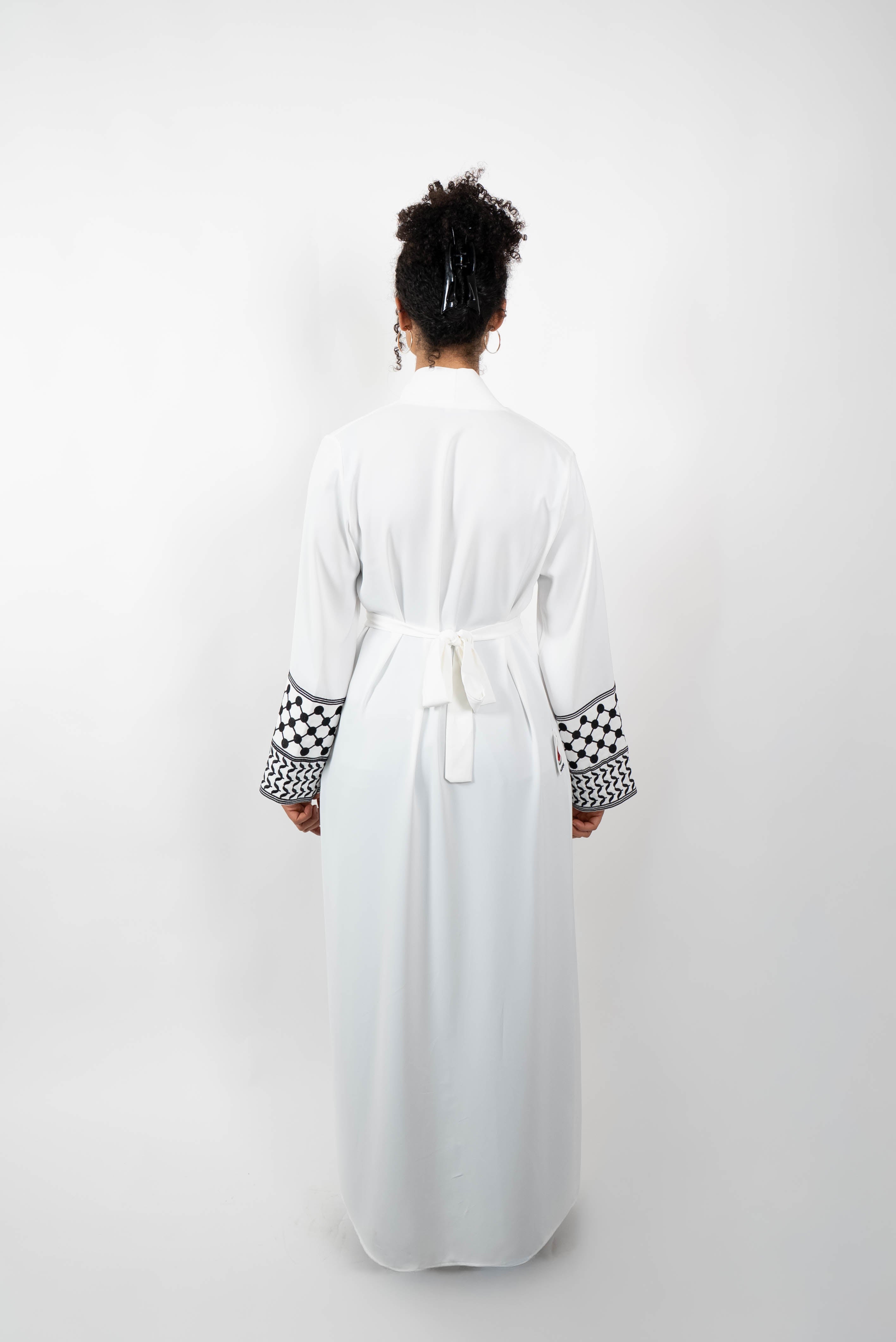 Rückenansicht einer Frau in einer langen, weißen Abaya, die mit einem Gürtel im Rücken geschlossen ist. Die Ärmelabschlüsse sind mit einem auffälligen schwarz-weißen Muster, das Kufiya-ähnlichen Mustern nachempfunden ist, verziert.