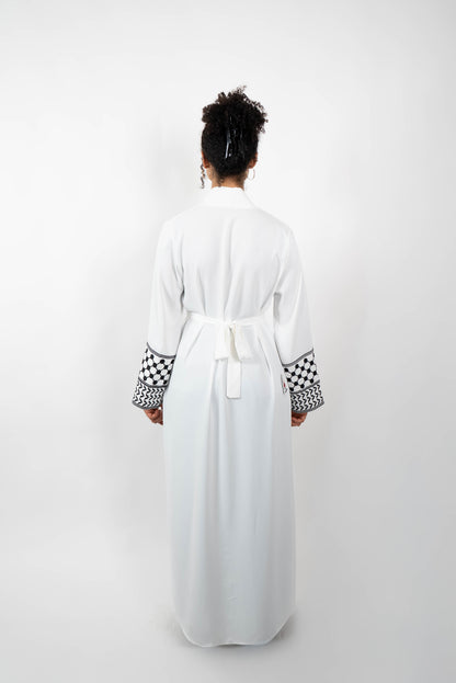 Rückenansicht einer Frau in einer langen, weißen Abaya, die mit einem Gürtel im Rücken geschlossen ist. Die Ärmelabschlüsse sind mit einem auffälligen schwarz-weißen Muster, das Kufiya-ähnlichen Mustern nachempfunden ist, verziert.