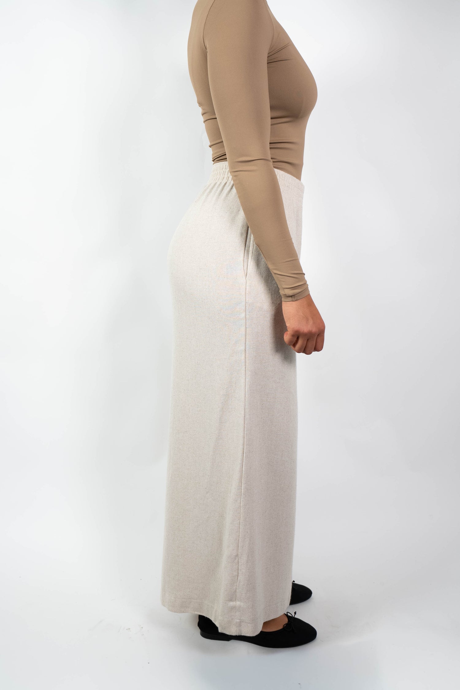 Seitenansicht einer Frau in einer cremefarbenen, weiten Marlene-Hose mit hohem, elastischem Bund und einem eng anliegenden, beige/braunen Oberteil.