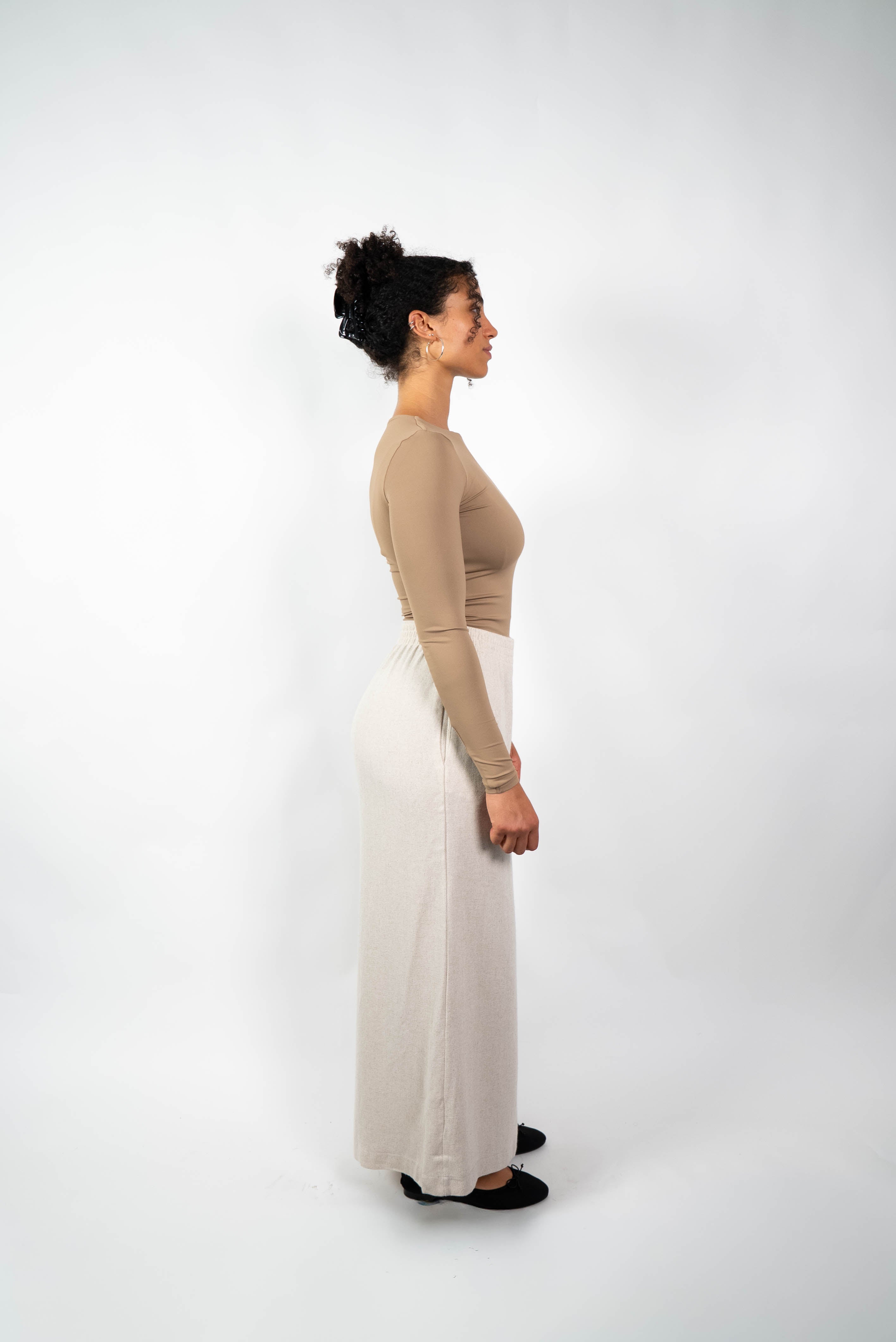 Seitenansicht einer Frau in einer cremefarbenen, weiten Marlene-Hose aus weichem Material und einem eng anliegenden, beige/braunen Langarmshirt.
