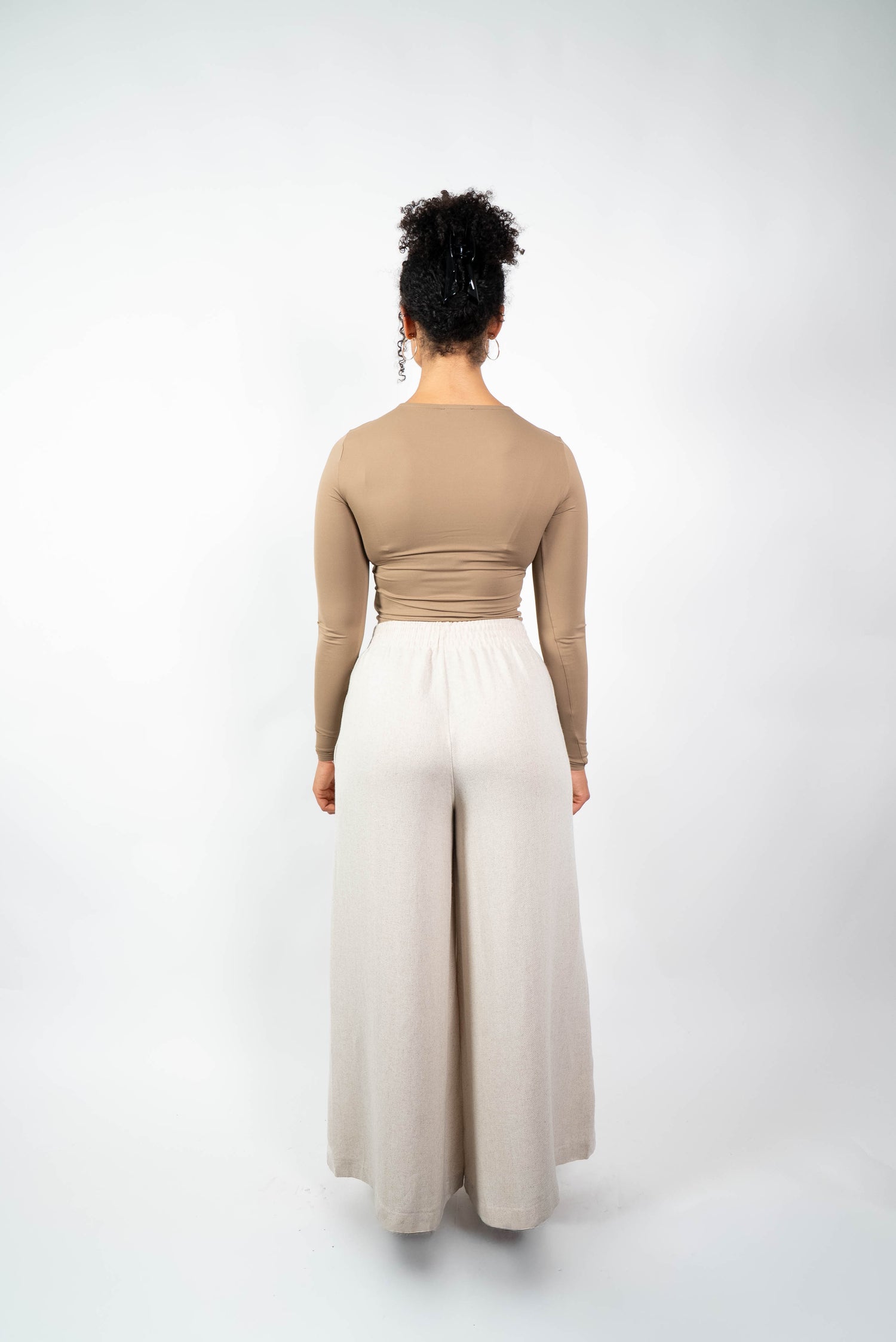 Rückenansicht einer Frau in einer cremefarbenen, weiten Marlene-Hose mit elastischem, gerafftem Bund und einem eng anliegenden, beige/braunen Oberteil.